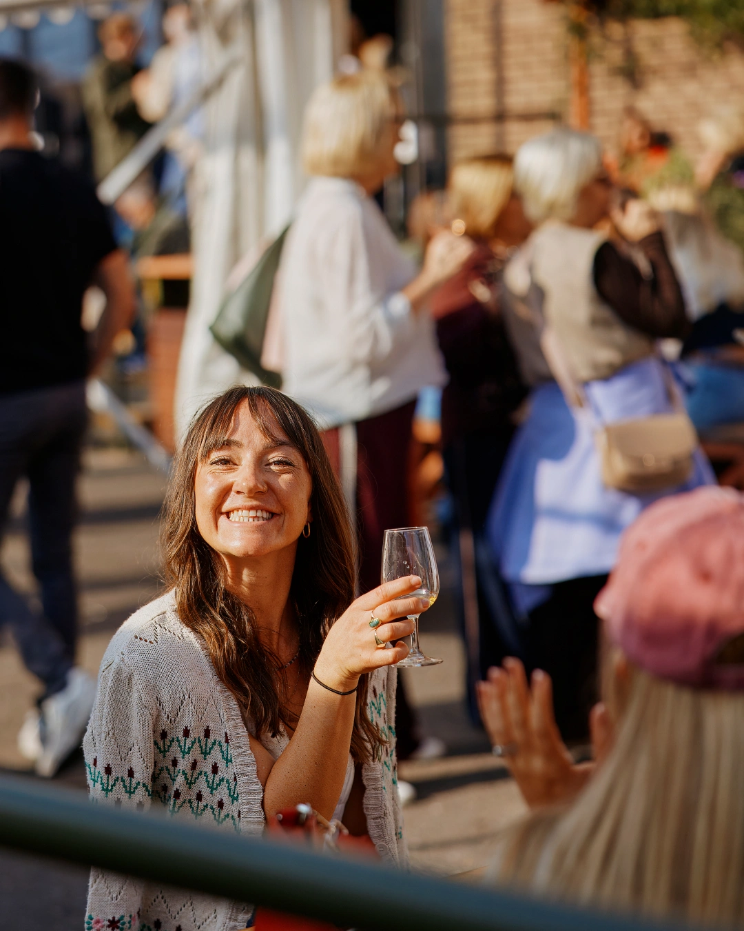 A smiling woman in a patterned cardigan holds a wine glass while sitting outdoors with other people.