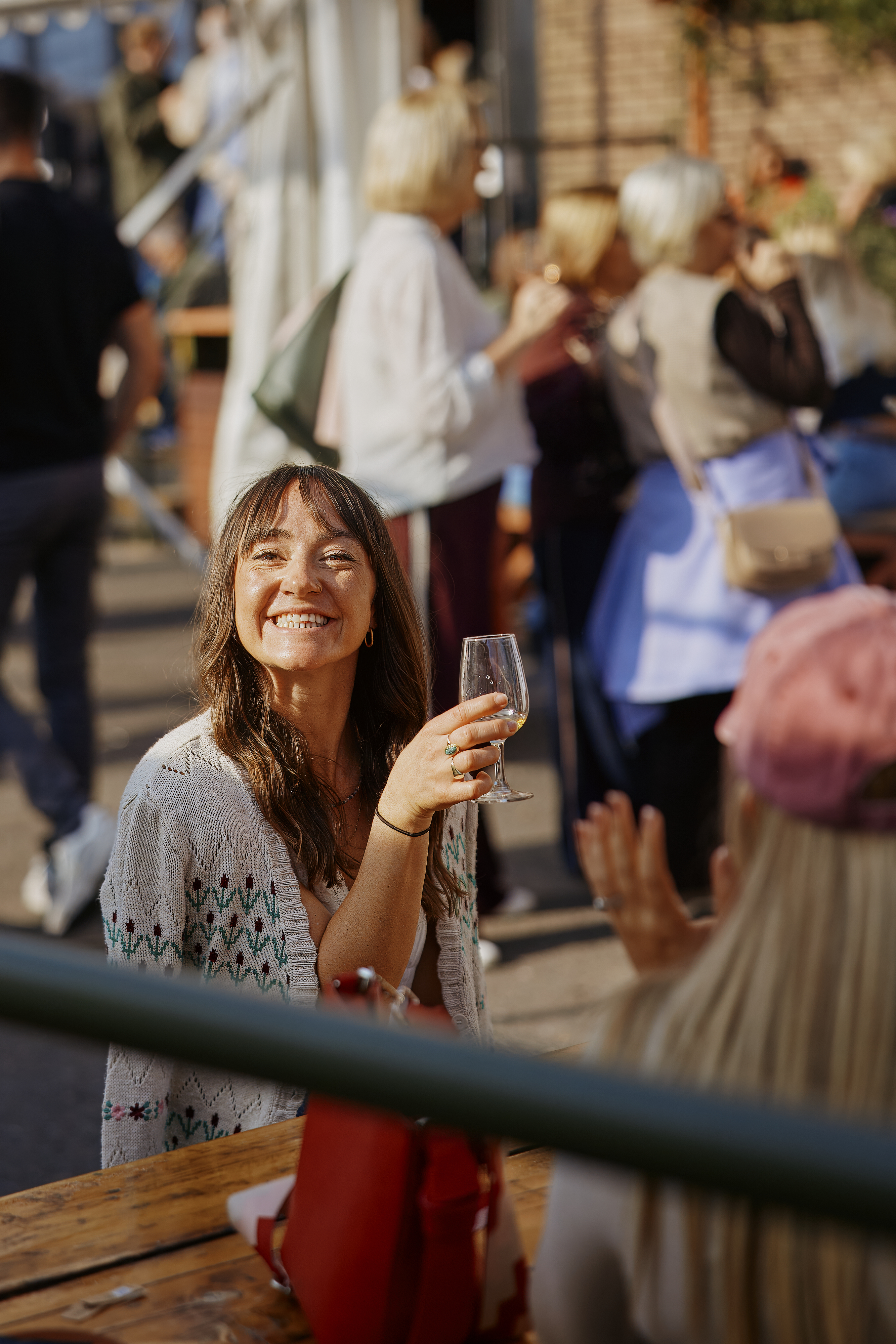Woman smiling outside, holding a glass, with people in the background.