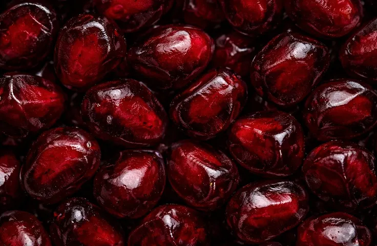 A close-up, macro photograph of glistening, deep red pomegranate seeds packed together.