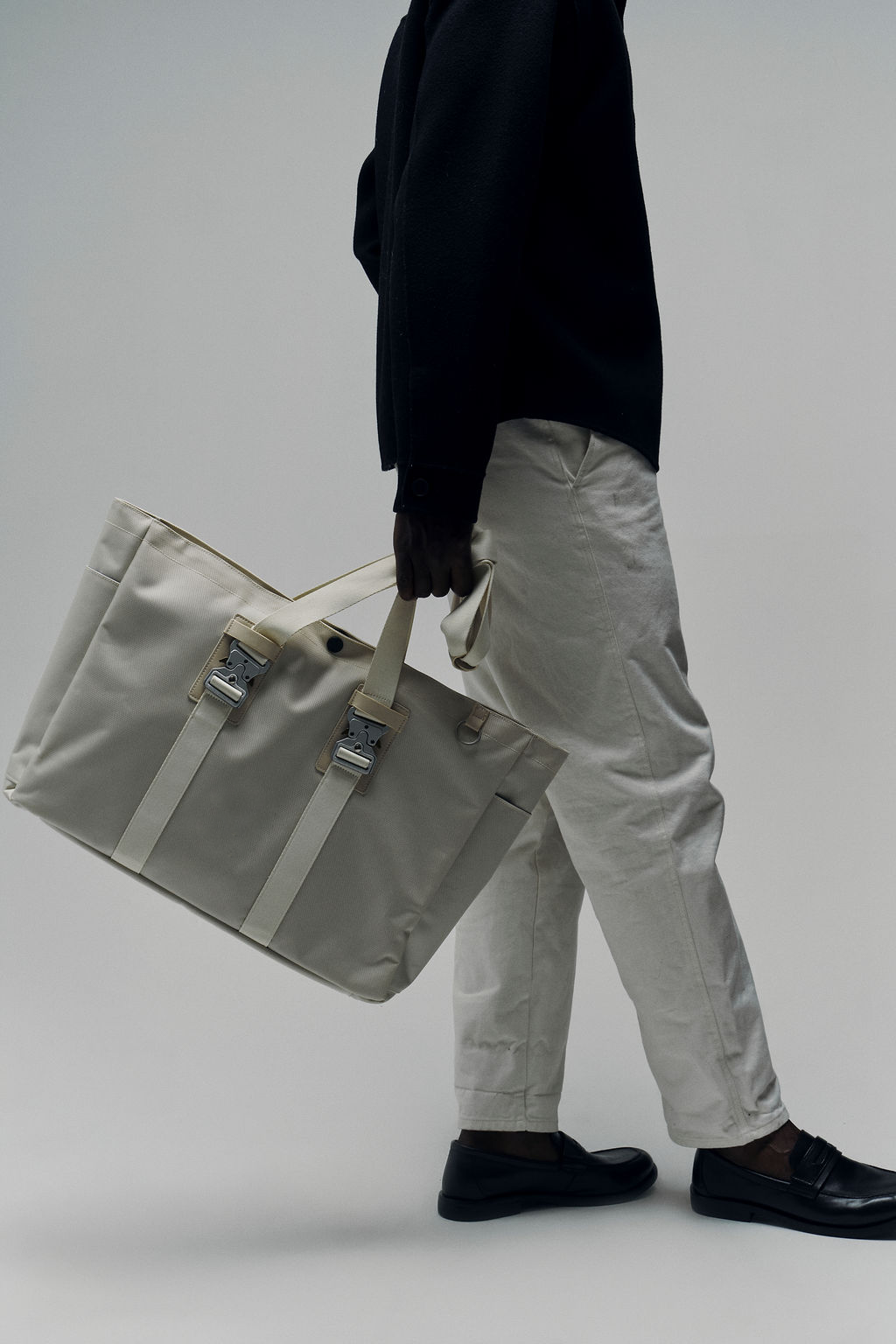 Person holding a large beige bag, wearing white pants and black shoes.