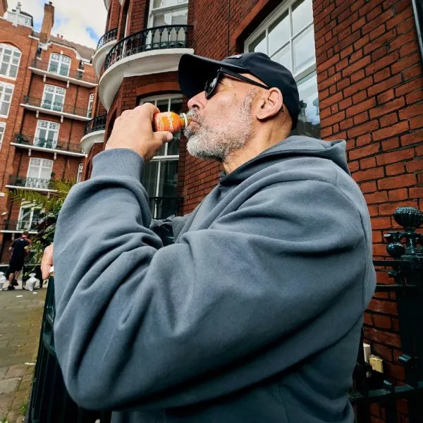 A man with a grey beard wearing a hoodie and cap drinks from a small bottle outdoors.