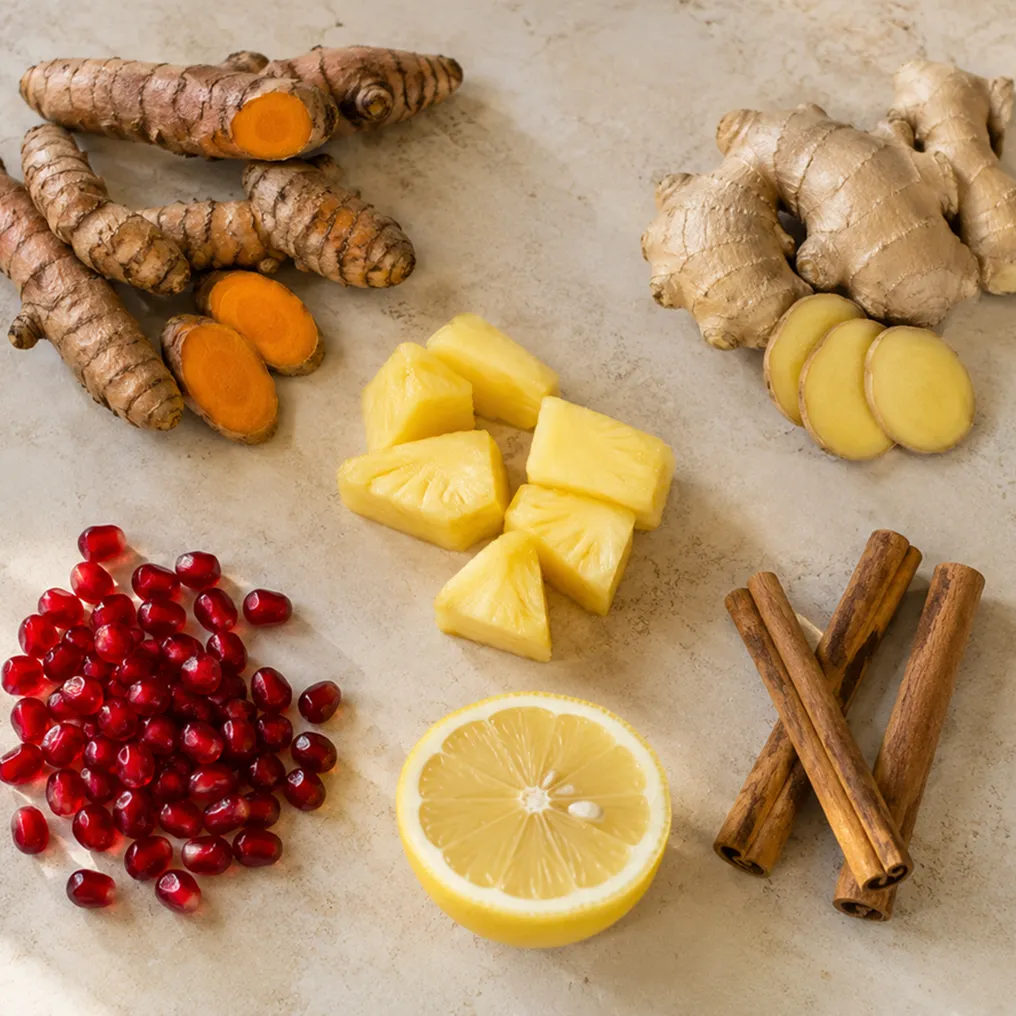 Turmeric, ginger, pineapple, pomegranate seeds, lemon, and cinnamon sticks arranged on a countertop.