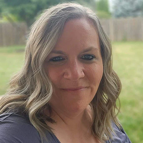A headshot of a smiling woman with wavy blonde hair, taken outdoors in a backyard.