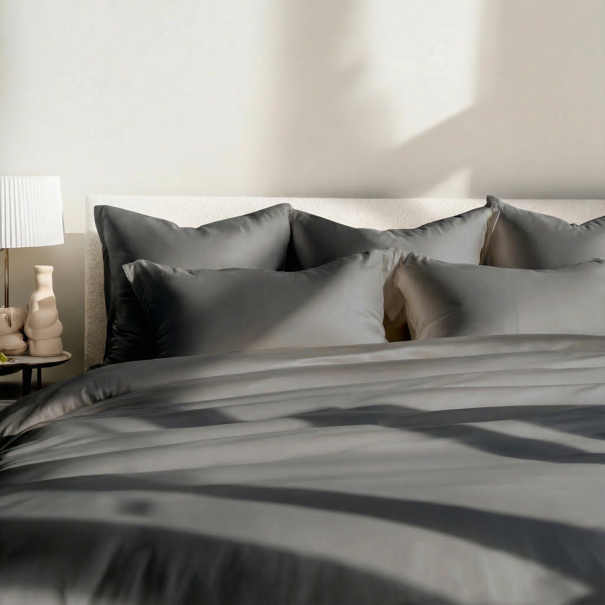 A sunlit bed with plush white pillows and a smooth duvet, next to a nightstand with a lamp.