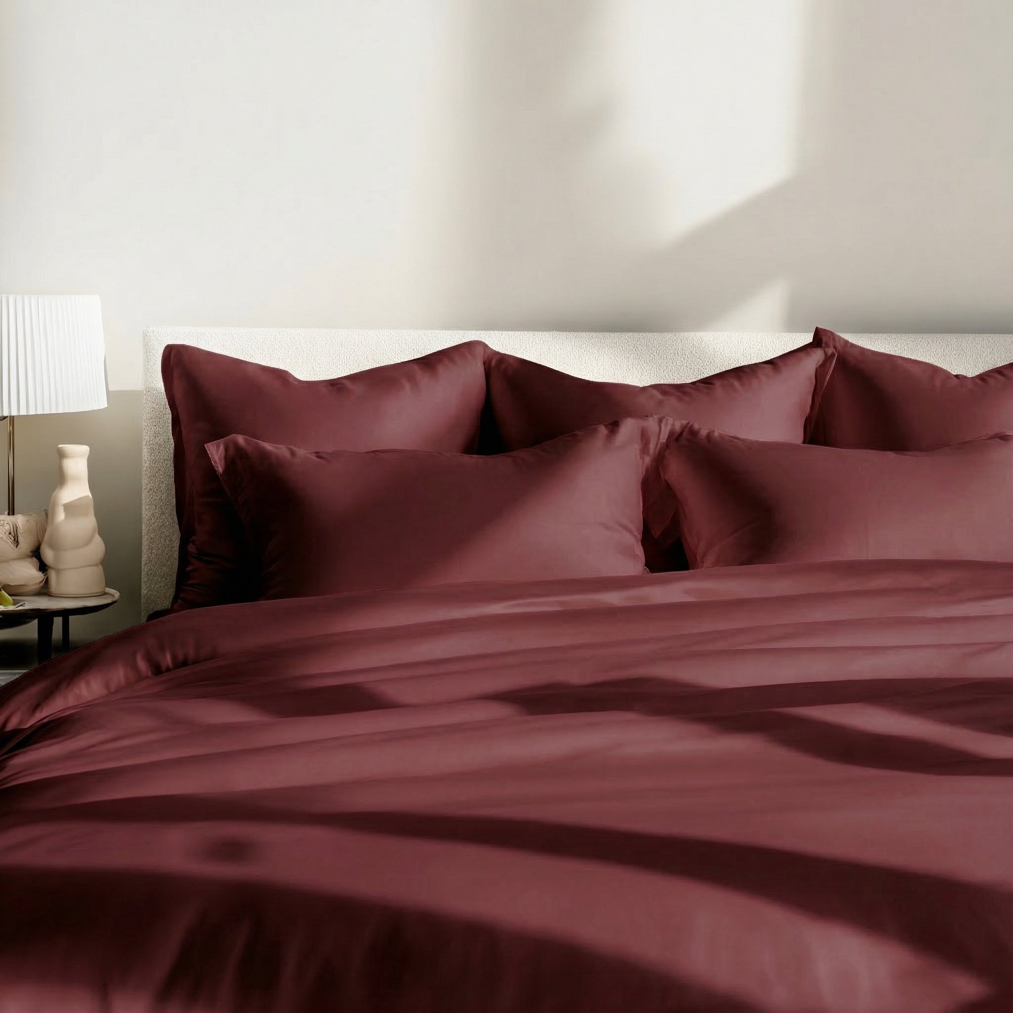 A close-up of a bed with deep red sateen bedding next to a nightstand with a lamp.