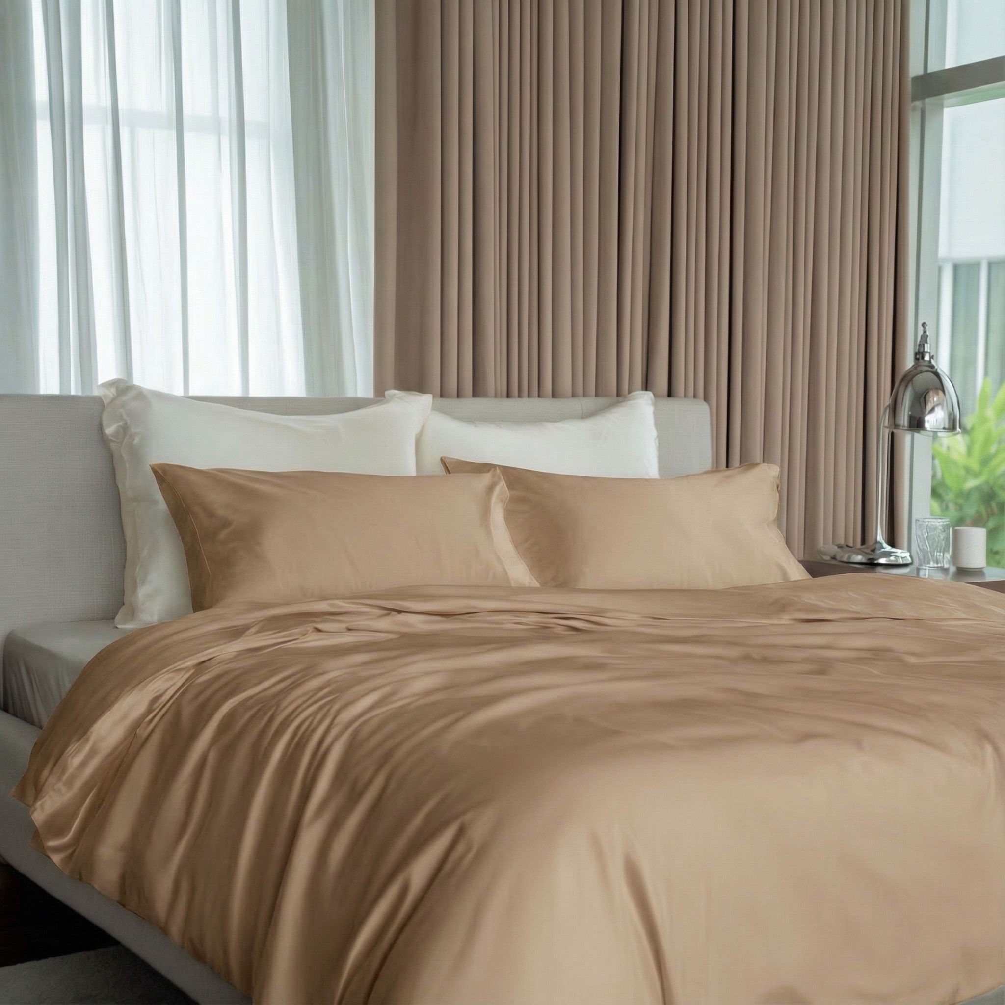 A neatly made bed with silky, light brown bedding in front of a window with curtains.