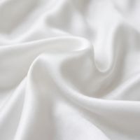 A sunlit bed with plush white pillows and a smooth duvet, next to a nightstand with a lamp.