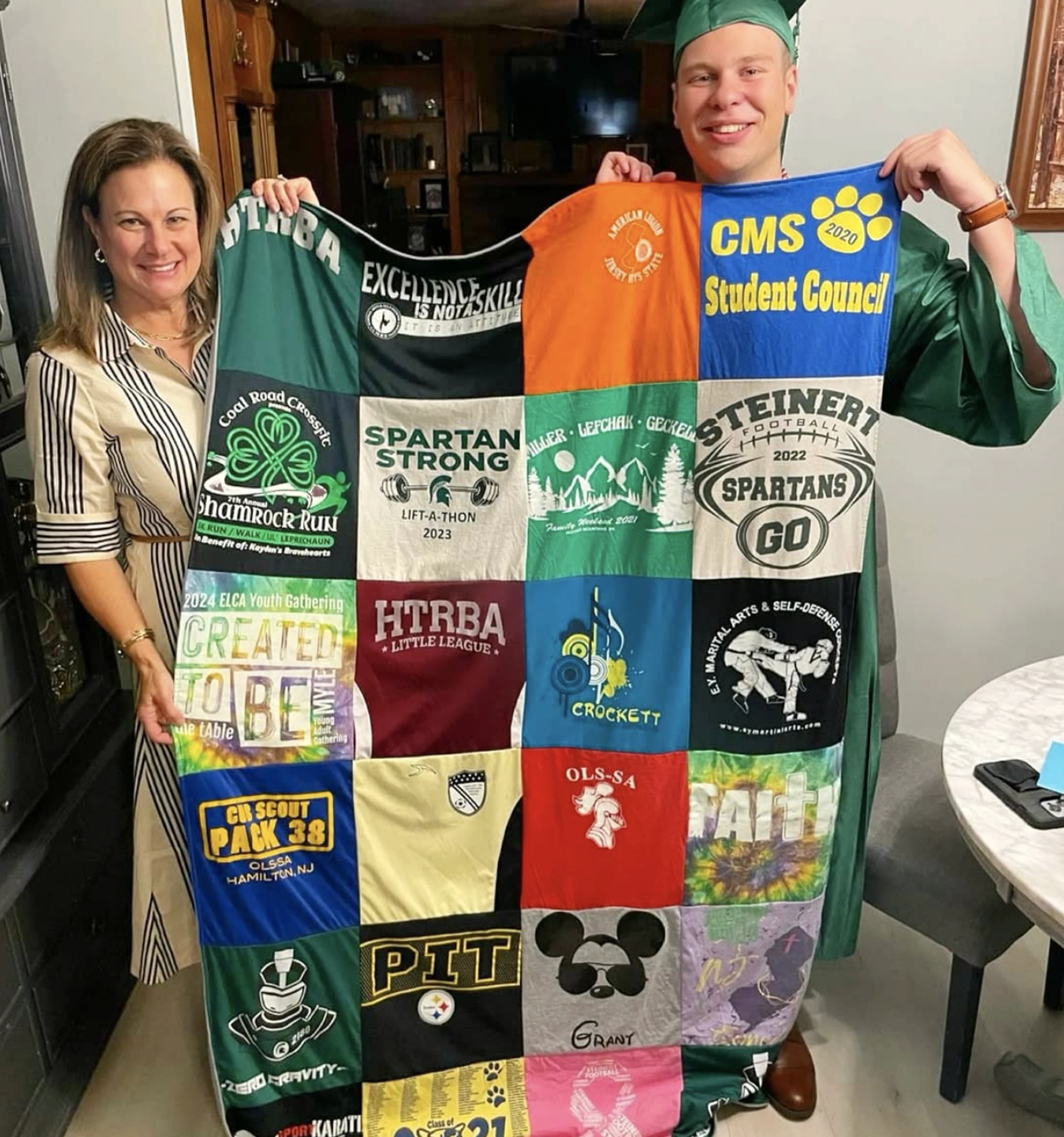 A woman and a young man in a graduation gown hold up a t-shirt quilt.