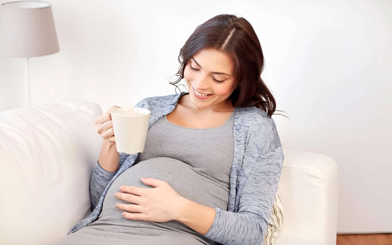 Pregnant woman sitting on a couch, holding a mug and smiling.