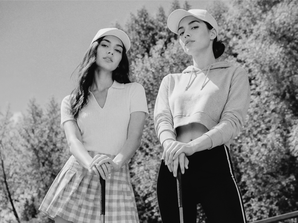 A low-angle, black-and-white photo of two women in caps holding golf clubs in front of trees.