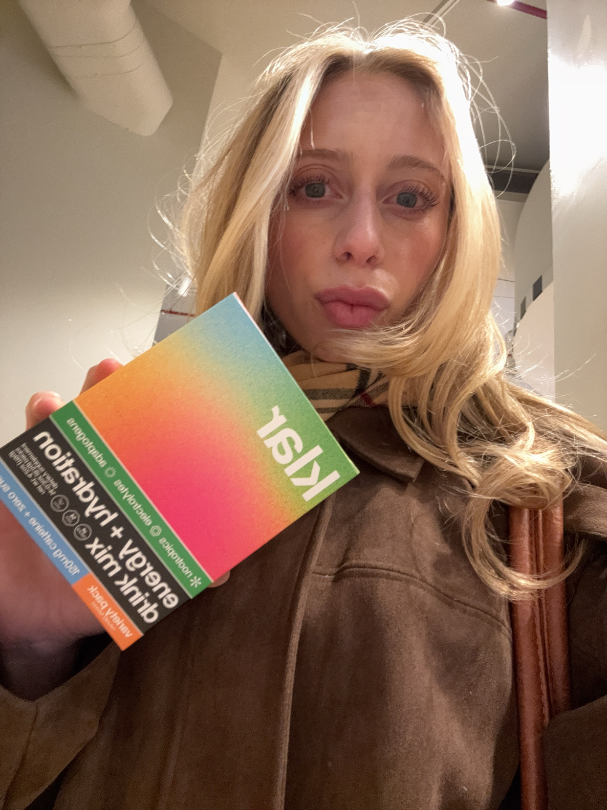 A blonde woman in a brown jacket takes a selfie while holding up a colorful box.