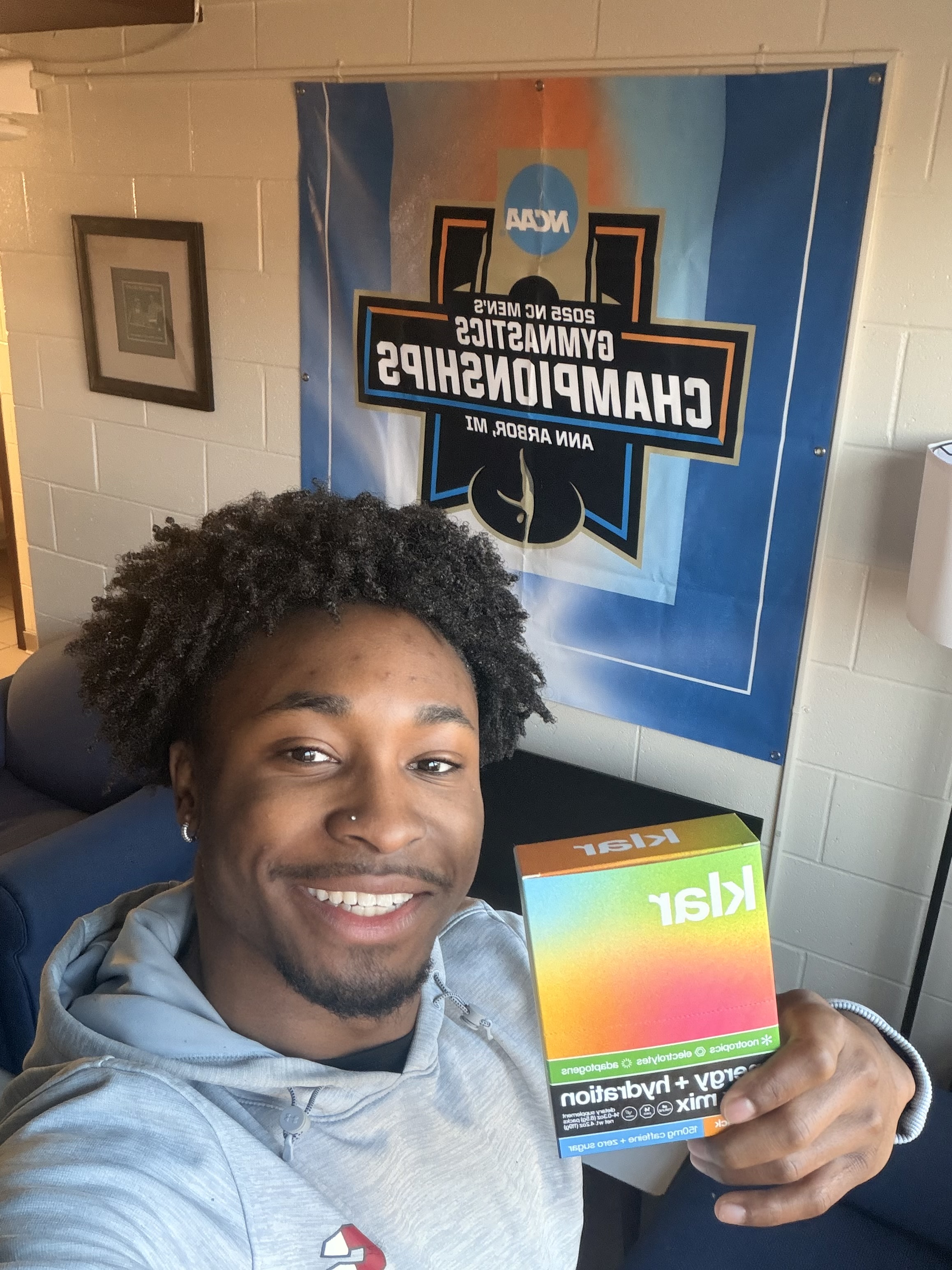 A smiling young man in a grey hoodie holds up a colorful box in front of a gymnastics banner.
