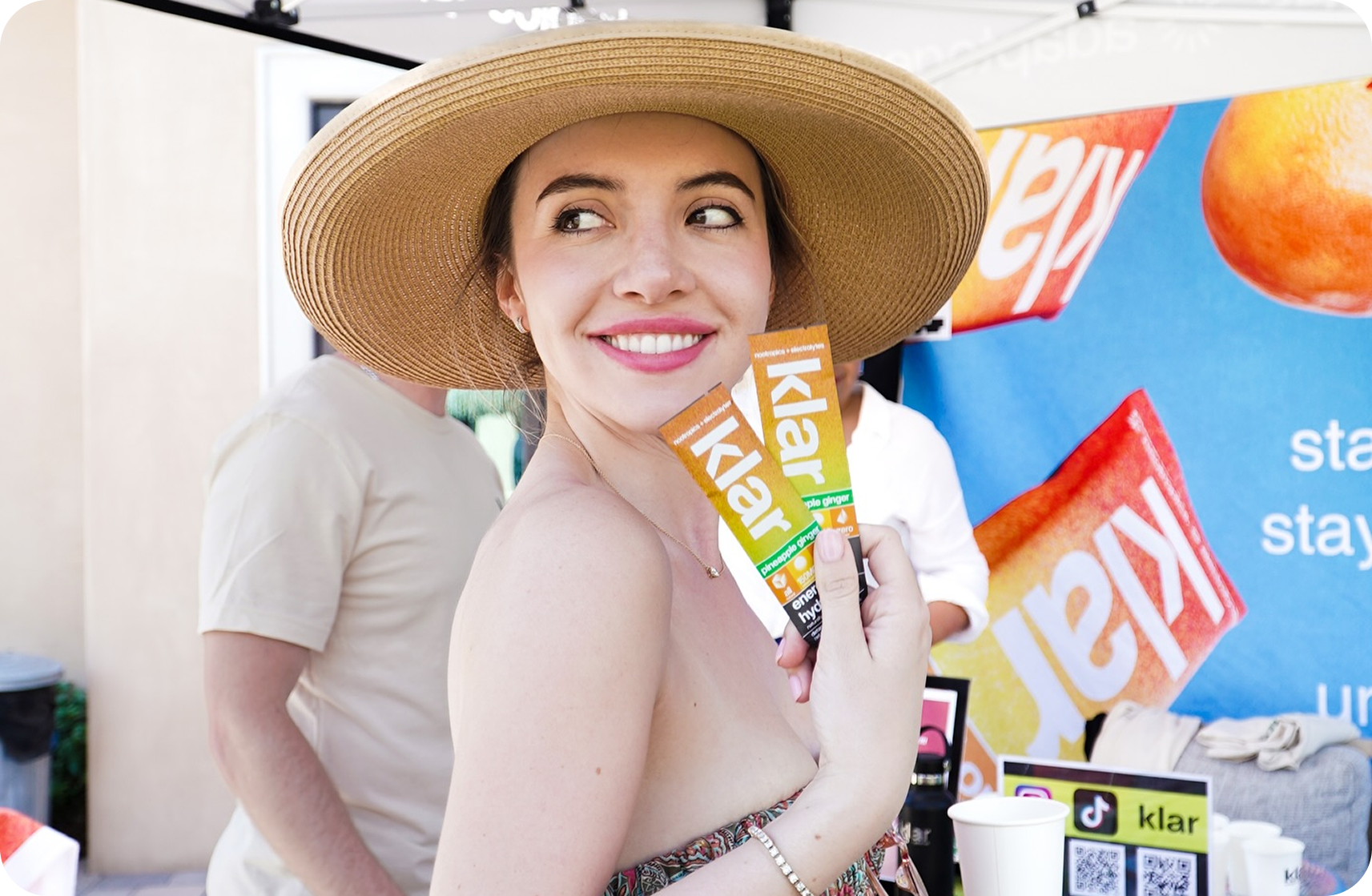 Woman in a hat holding Klar energy drink packets, smiling at an outdoor event.