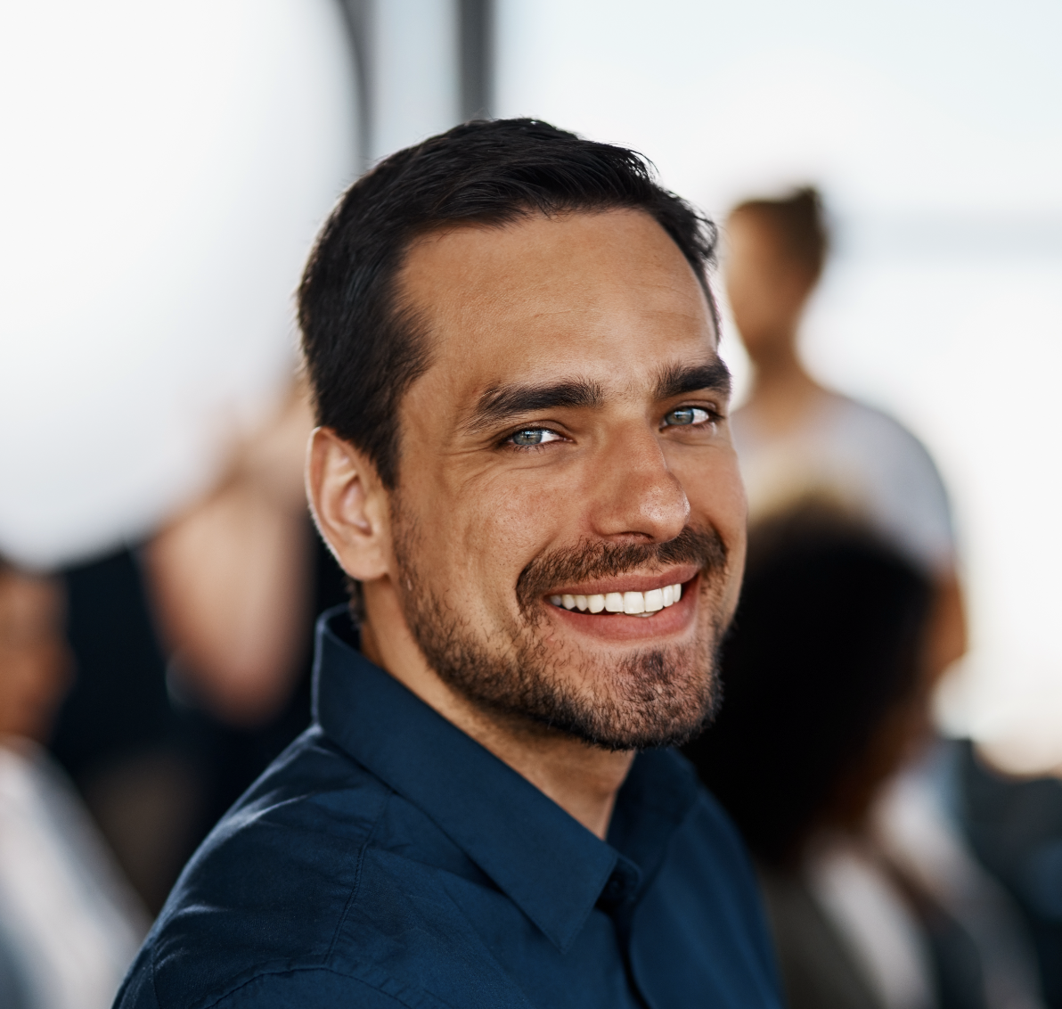 A close-up portrait of a smiling man with dark hair and a short beard, wearing a blue shirt.