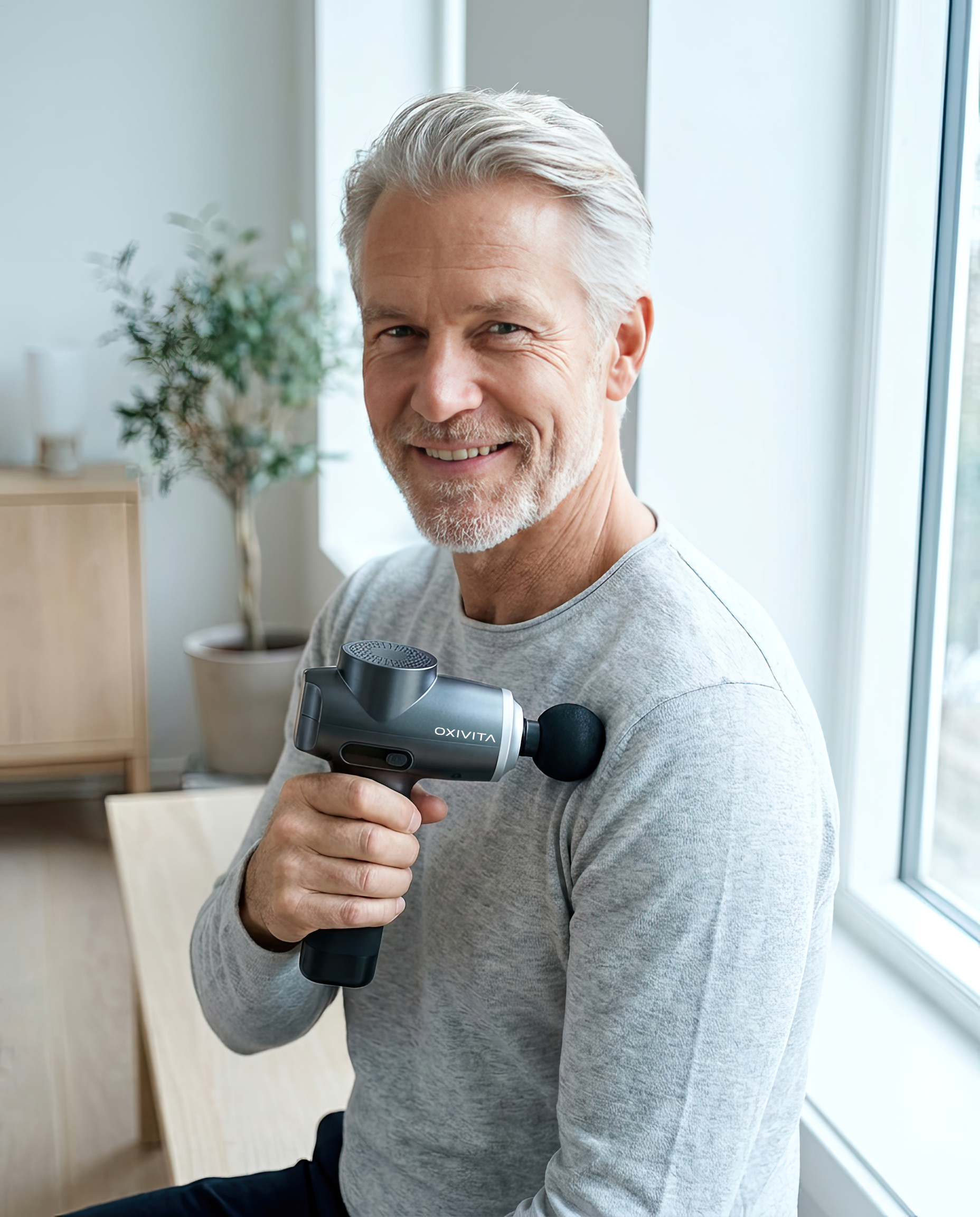 A smiling, grey-haired man holds a handheld massage gun to his shoulder while looking at the camera.