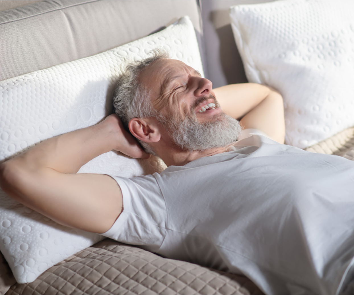 Smiling man with a beard relaxing on a bed with white pillows.