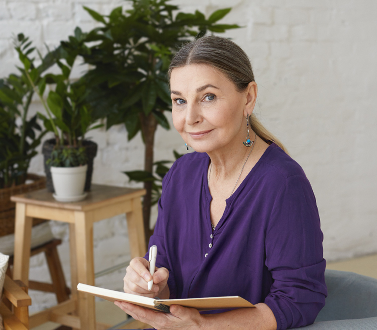 Woman in purple shirt writing in a notebook, surrounded by plants.