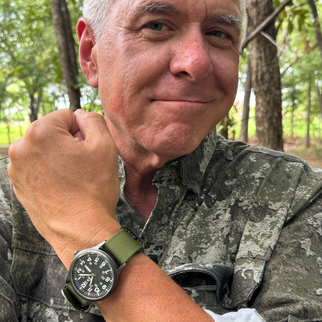 Person in camo shirt showing a wristwatch with green band, outdoors in a wooded area.