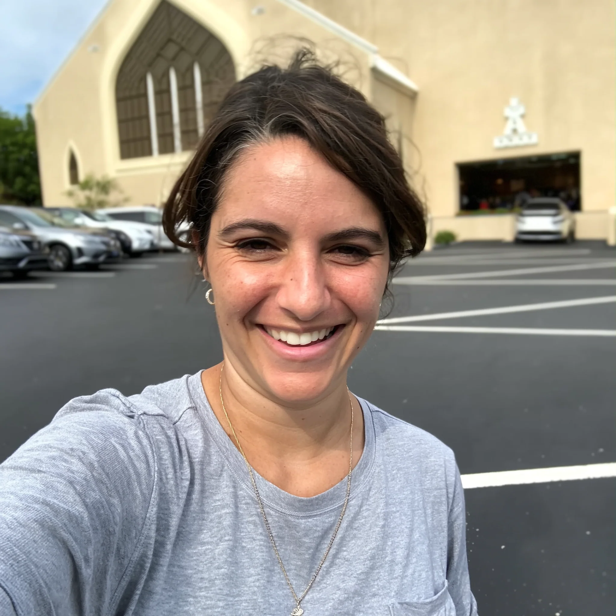 A woman with dark hair smiles for a selfie in a parking lot in front of a church.