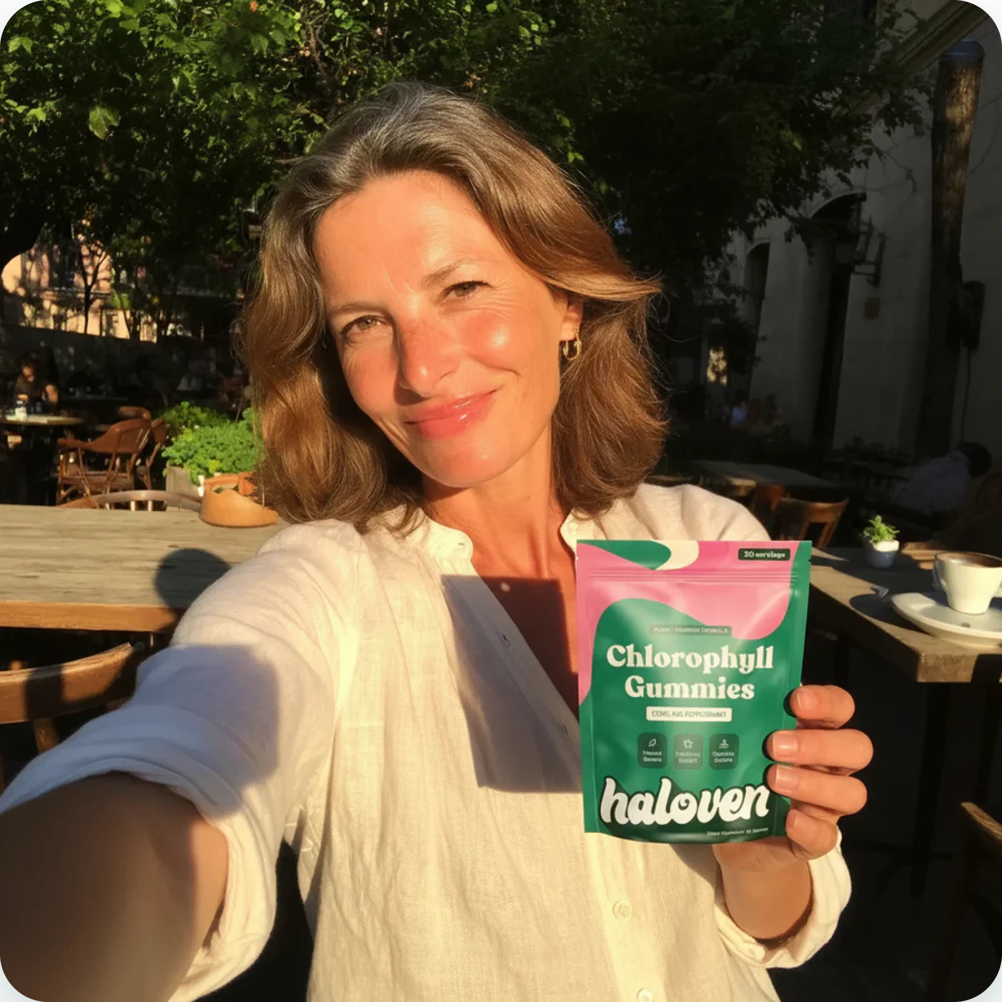 A woman smiles while holding a pouch of Haloven Chlorophyll Gummies at an outdoor cafe.