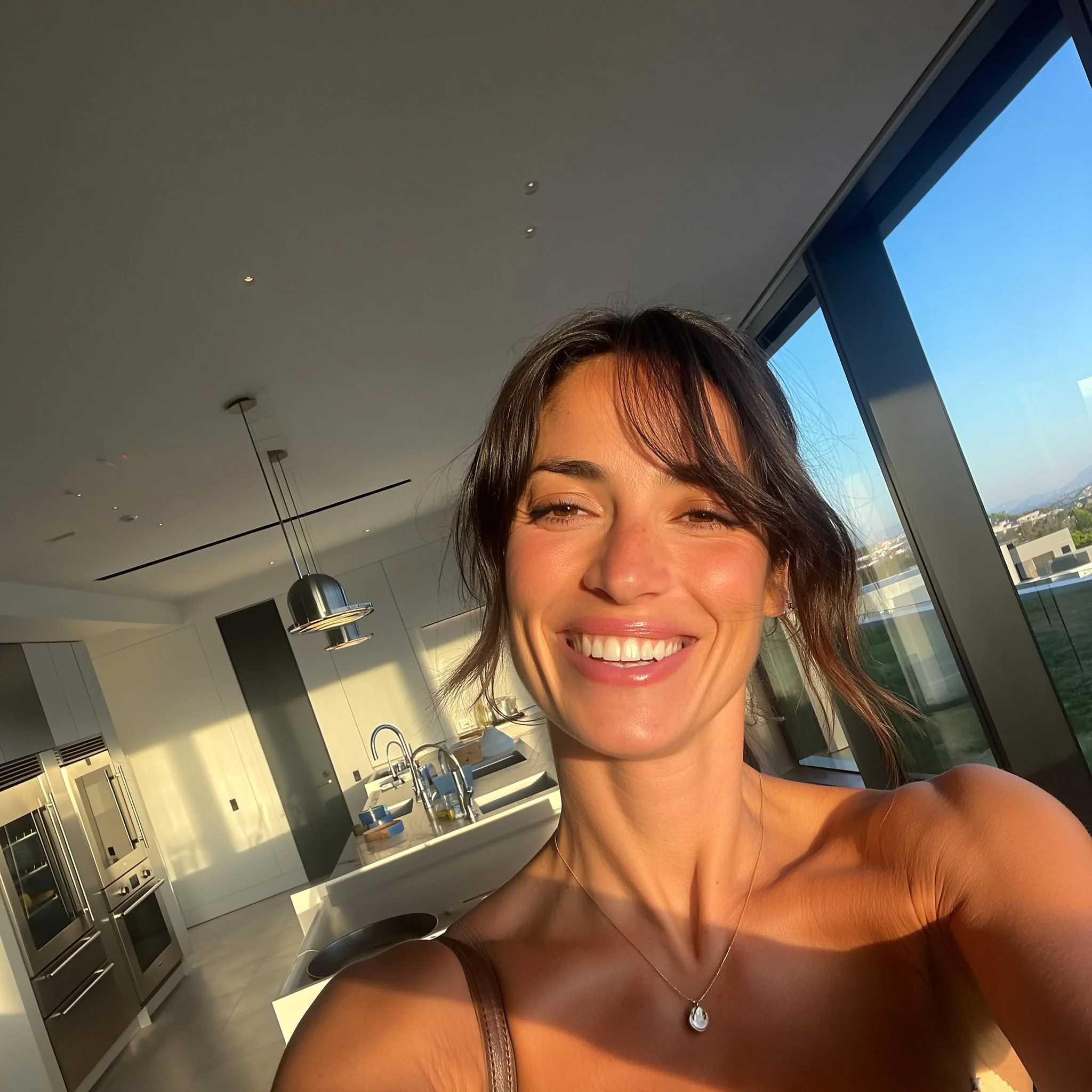 A smiling woman with dark hair takes a selfie in a modern, sunlit kitchen with large windows.