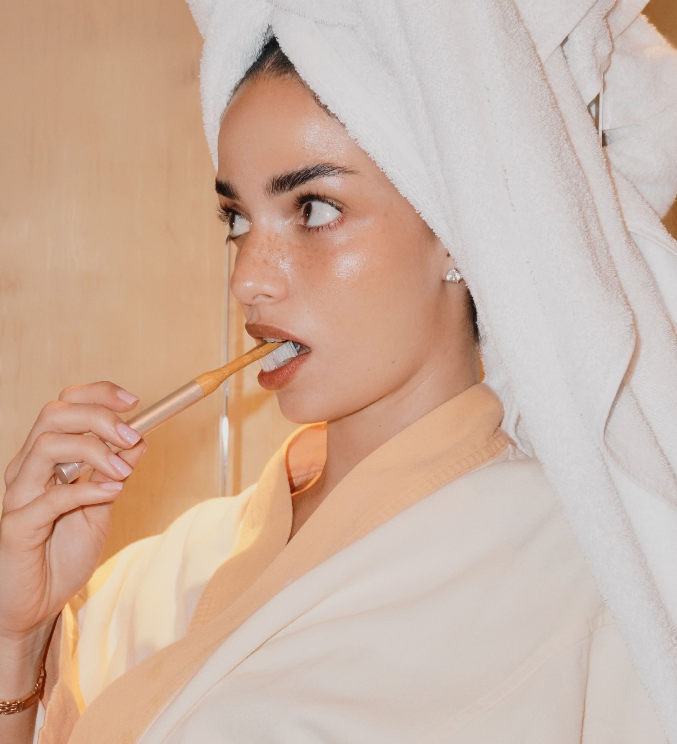A woman in a bathrobe with a towel on her head is brushing her teeth.
