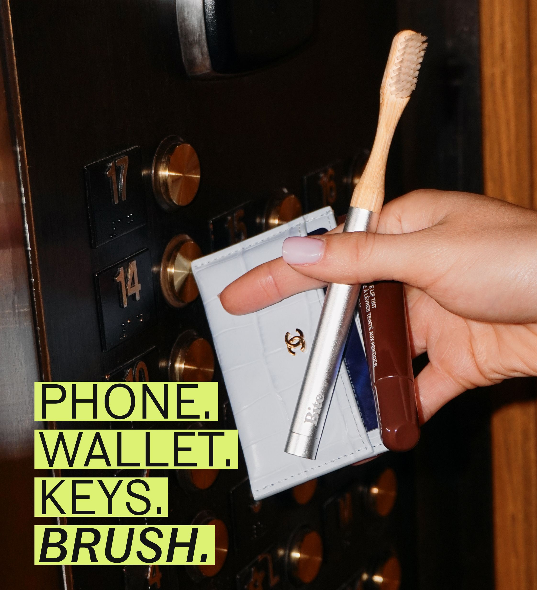 A hand holds a wallet, toothbrush, and lip tint in front of an elevator button panel.