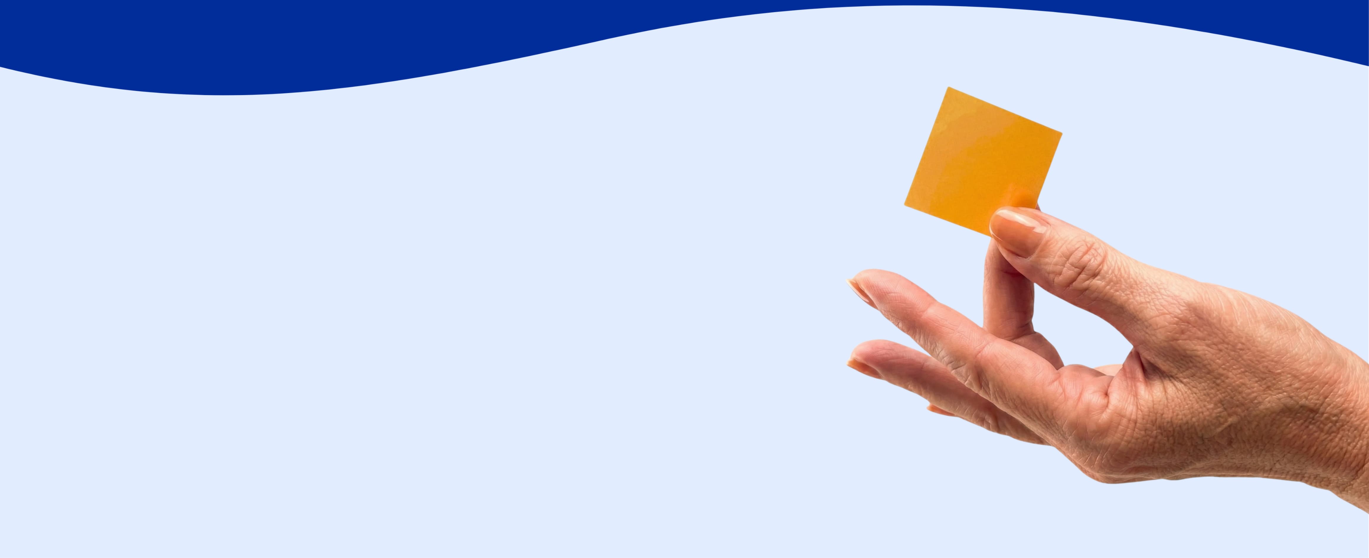 A person's hand holds a small orange square against a light blue background with a dark blue wave.