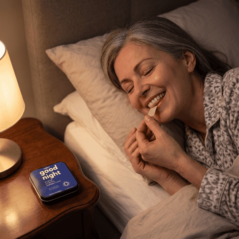 A smiling woman in bed applies a sleep strip to her tongue next to a lamp and product tin.