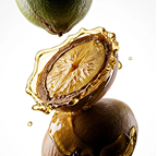 A stack of argan nuts, one split open, with golden oil splashing around it against a white background.