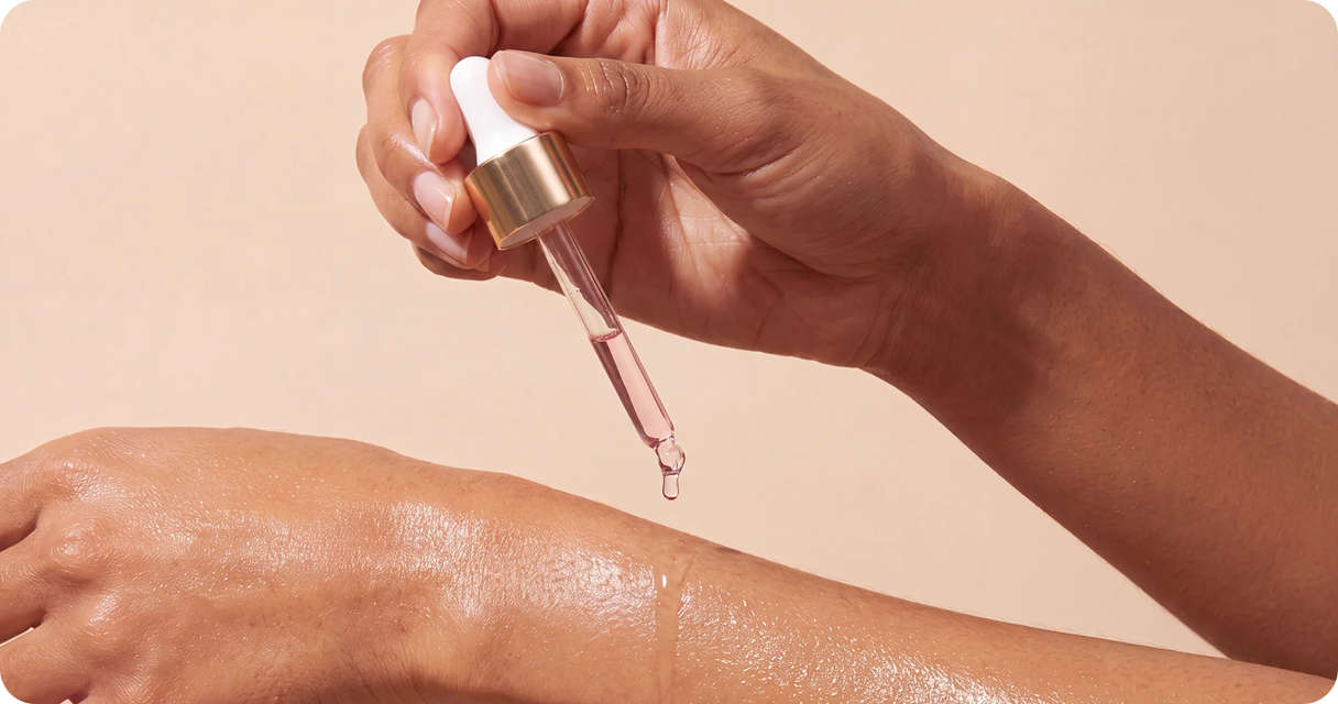 A person applies a drop of pink serum from a dropper onto the back of their hand.