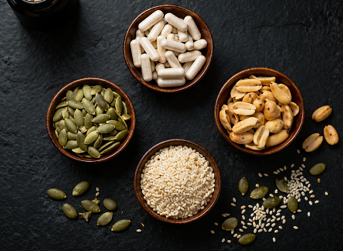 Four wooden bowls with pumpkin seeds, peanuts, sesame seeds, and vitamin capsules on a black slate background.