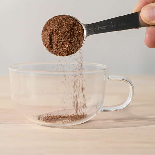 Spoon pouring ground coffee into a glass cup on a wooden surface.