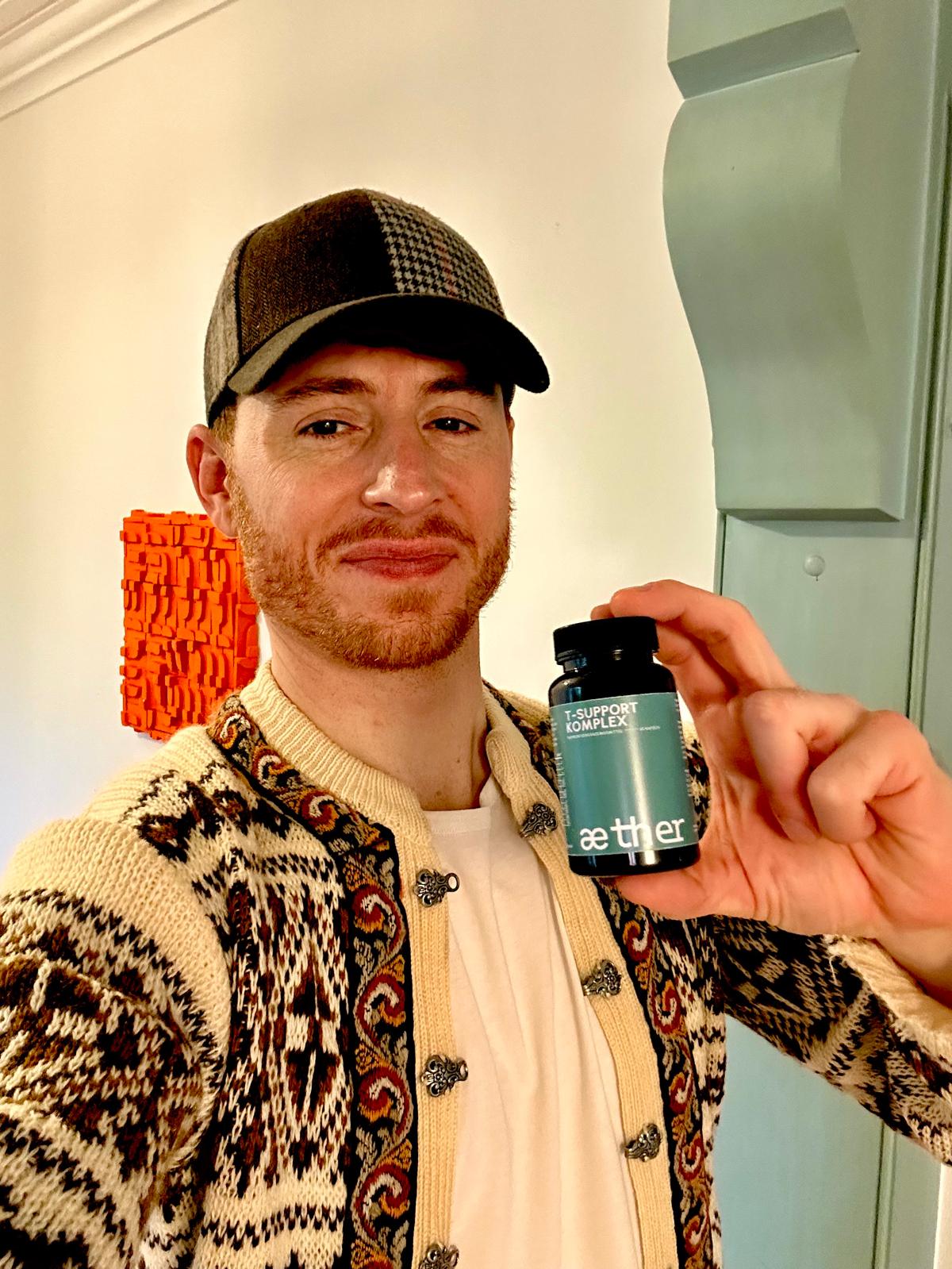 Person holding a bottle labeled 'T-Support Komplex', wearing a patterned cardigan and cap.
