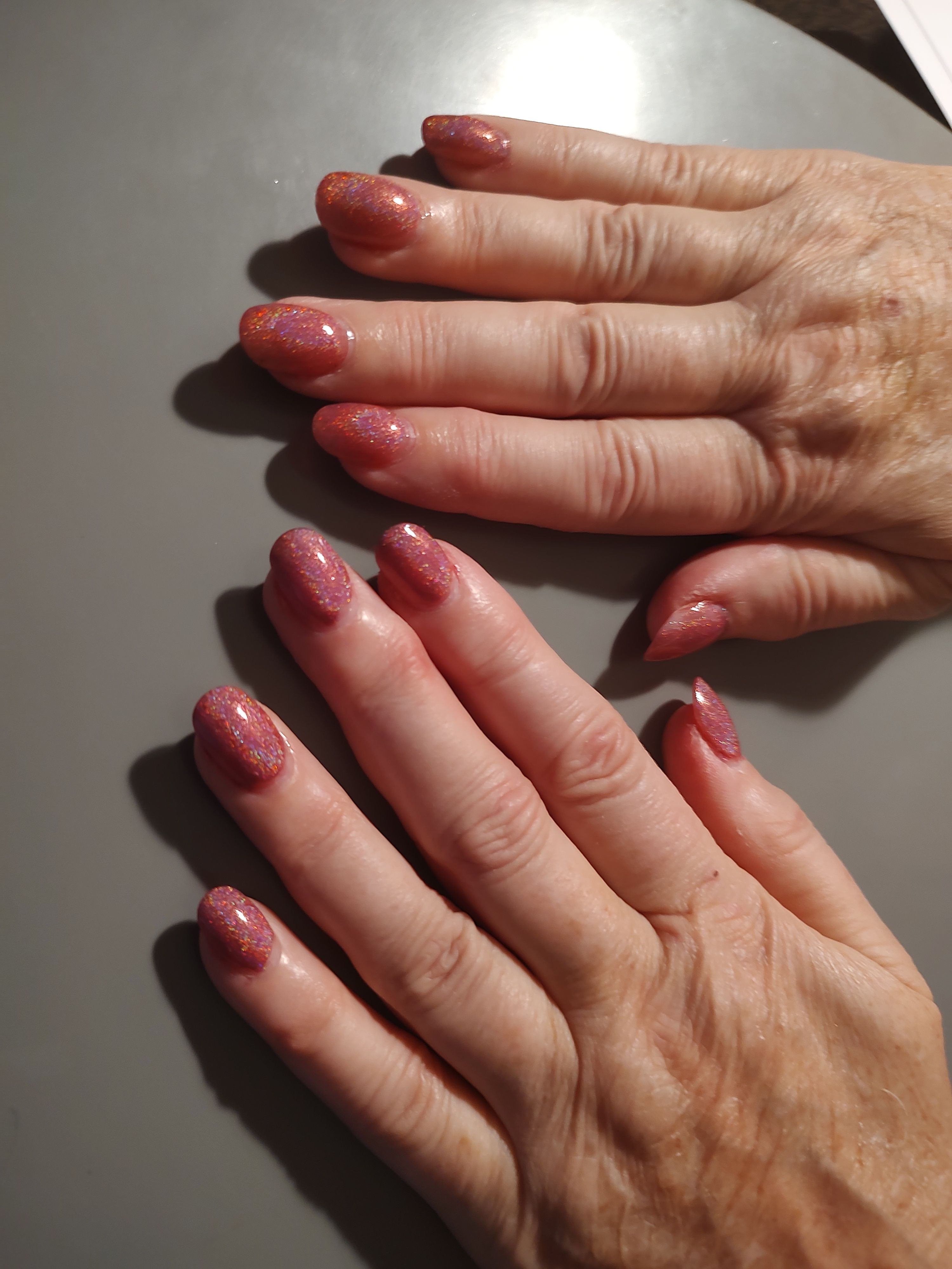 Two hands with glittery pink nail polish on a gray surface.