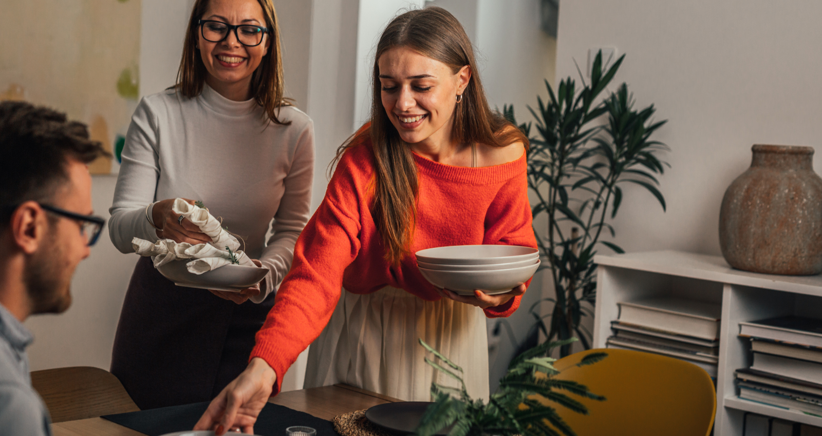 People setting up a dining table with plates and napkins, smiling and enjoying each other's company.