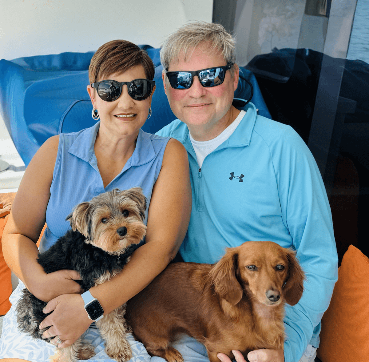 Two people sitting with two small dogs, wearing sunglasses, smiling.