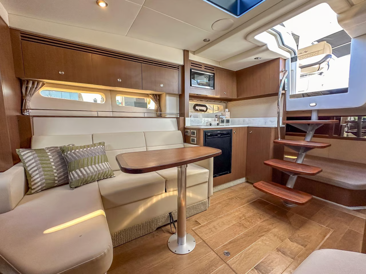 The interior of a boat cabin showing a seating area with a table, a kitchenette, and stairs.