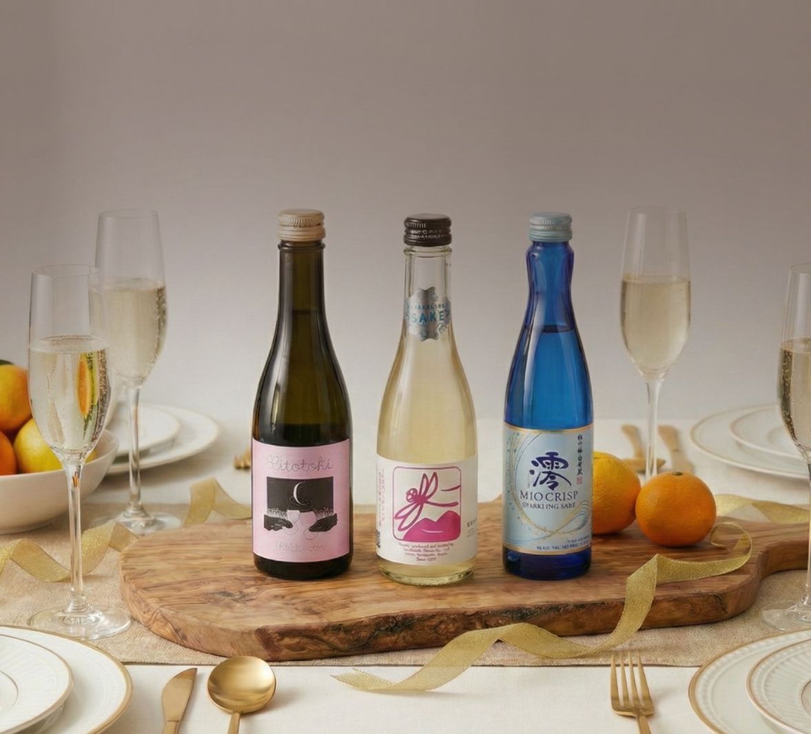 Three bottles of sparkling sake on a wooden board on a set table with champagne flutes and oranges.