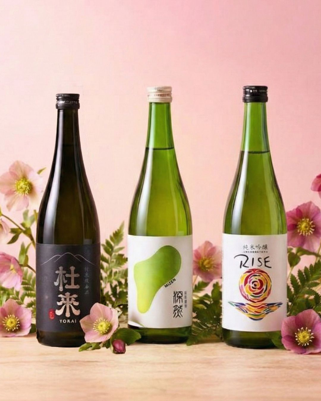 Three bottles of sake are arranged on a wooden surface surrounded by pink flowers against a pink background.