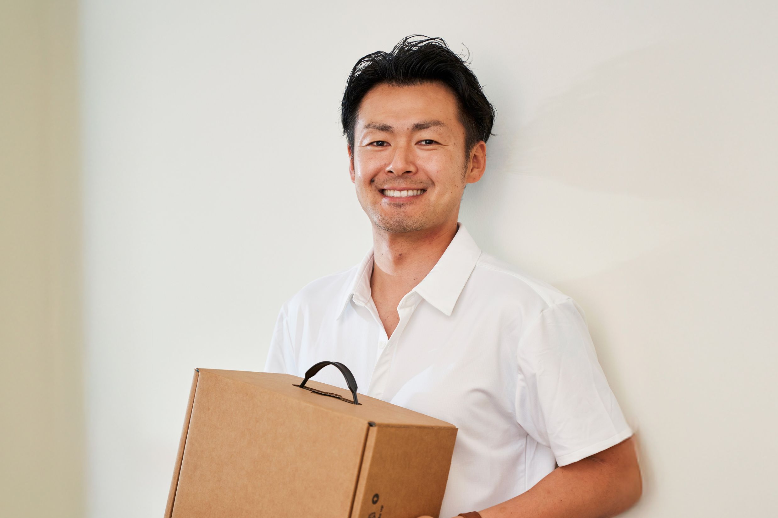 A smiling man in a white collared shirt holds a cardboard box while standing against a plain wall.