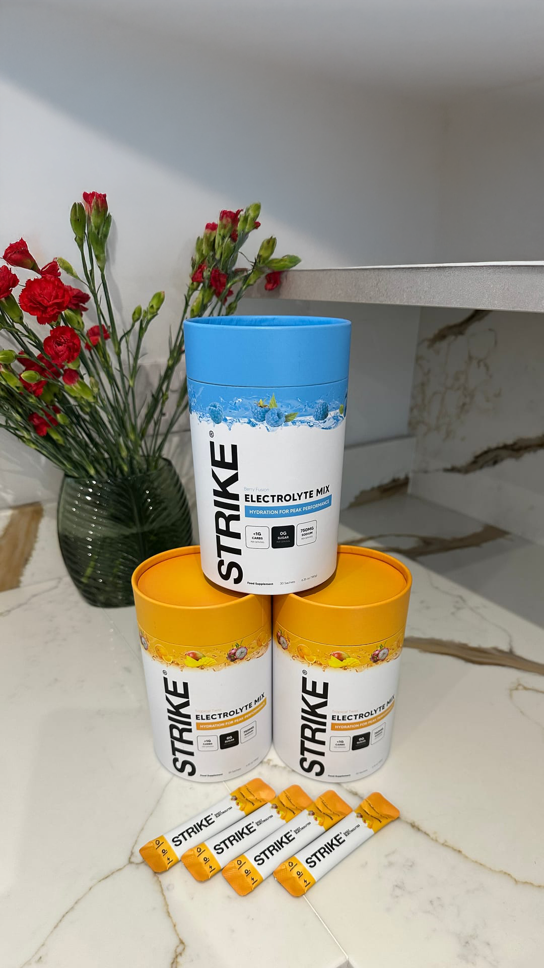 Three containers and packets of Strike Electrolyte Mix on a marble countertop with red flowers in the background.