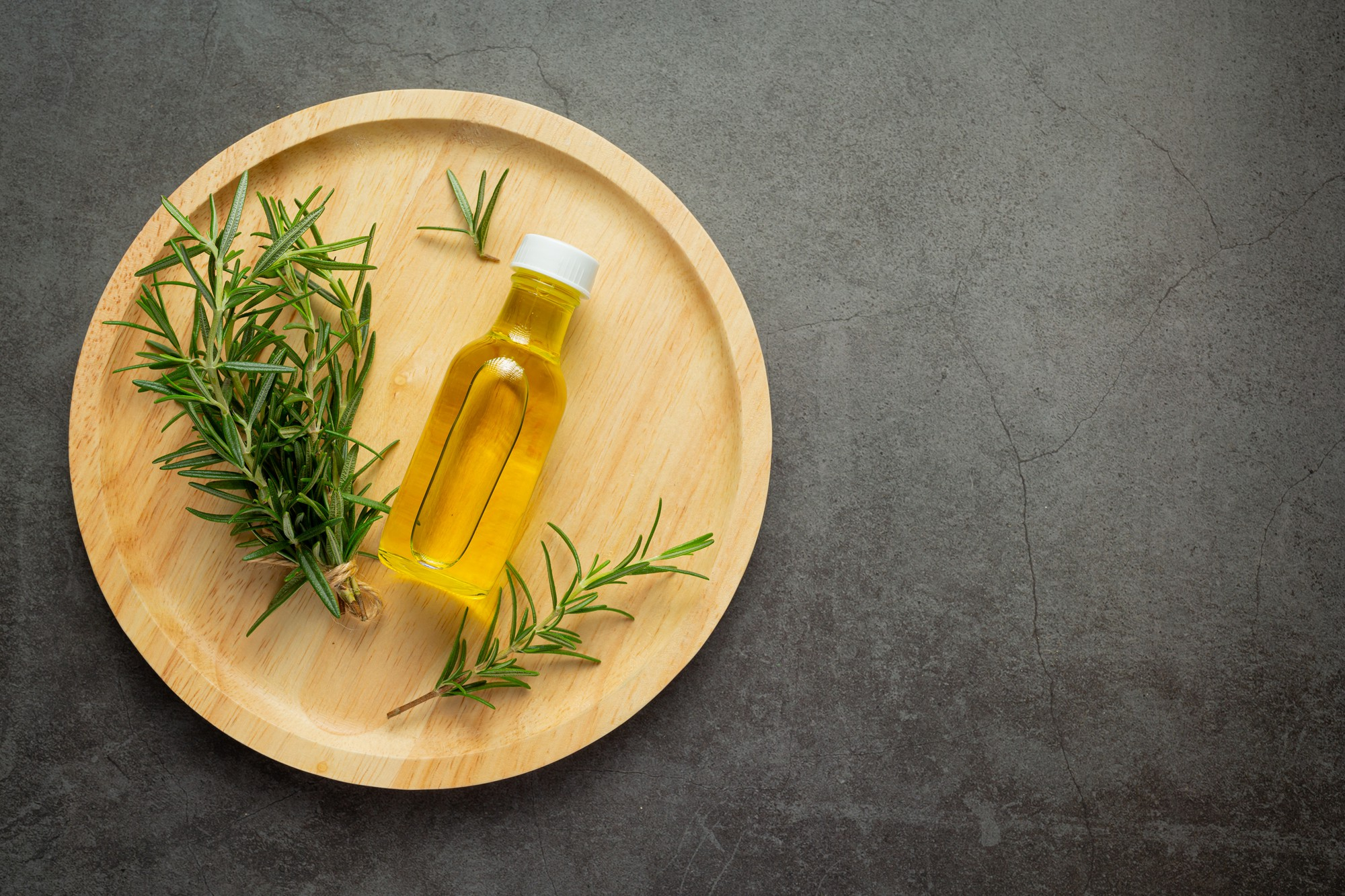 A top-down view of a bottle of oil and fresh rosemary sprigs on a round wooden plate.