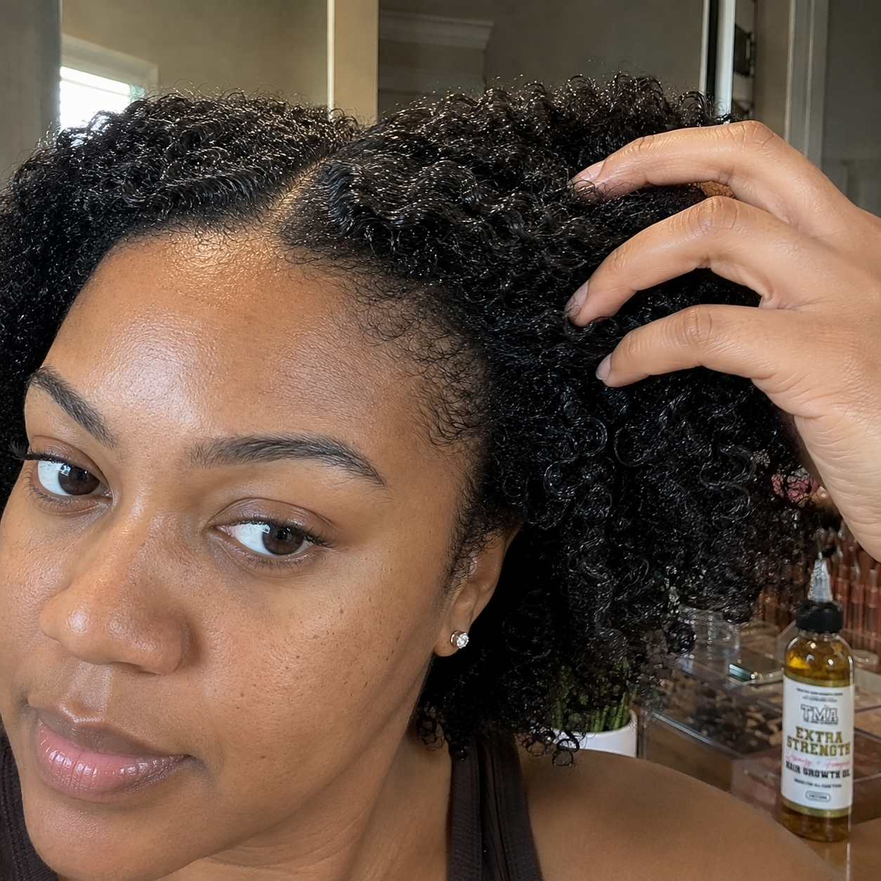 A woman runs her fingers through her curly hair, showing her scalp with a bottle of hair oil nearby.