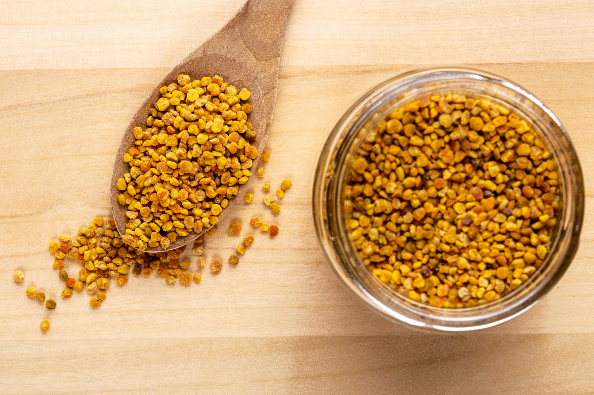 An overhead view of bee pollen granules in a glass jar and on a wooden spoon.