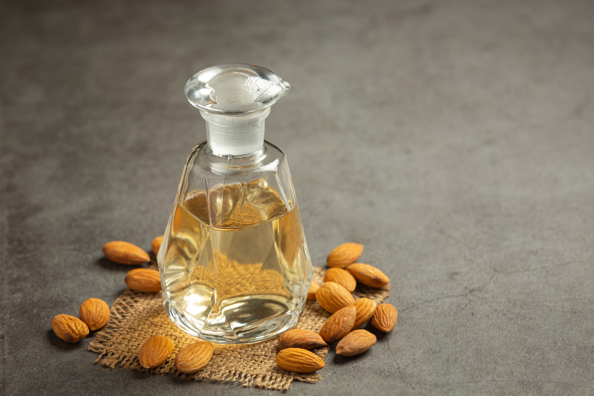 A glass bottle of golden oil sits on a burlap mat surrounded by almonds on a gray surface.