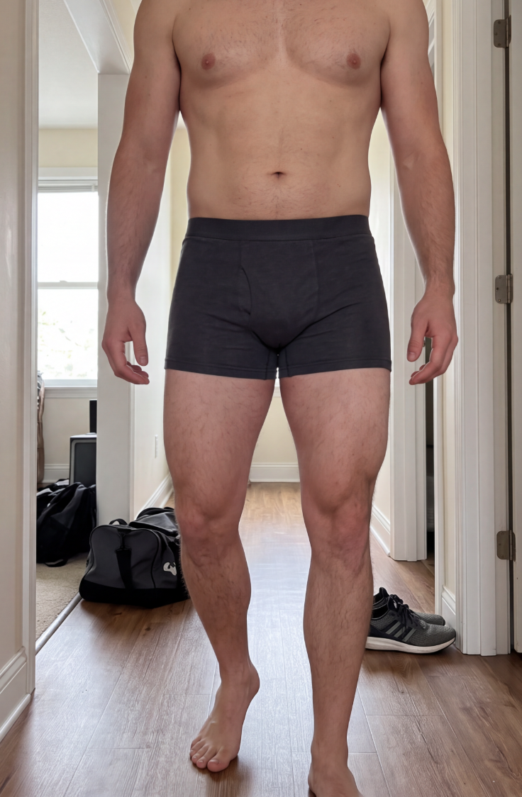 A muscular man wearing dark grey boxer briefs stands in a hallway.