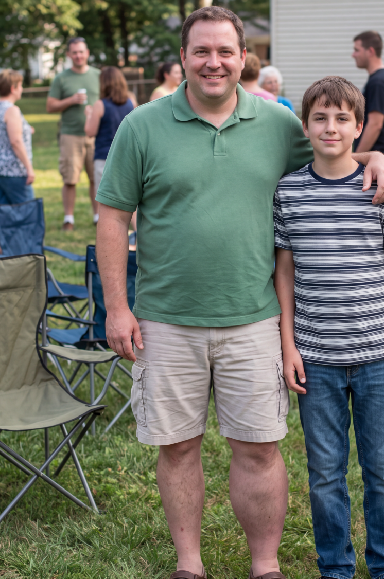 A smiling man in a green shirt poses with a boy in a striped shirt outdoors.