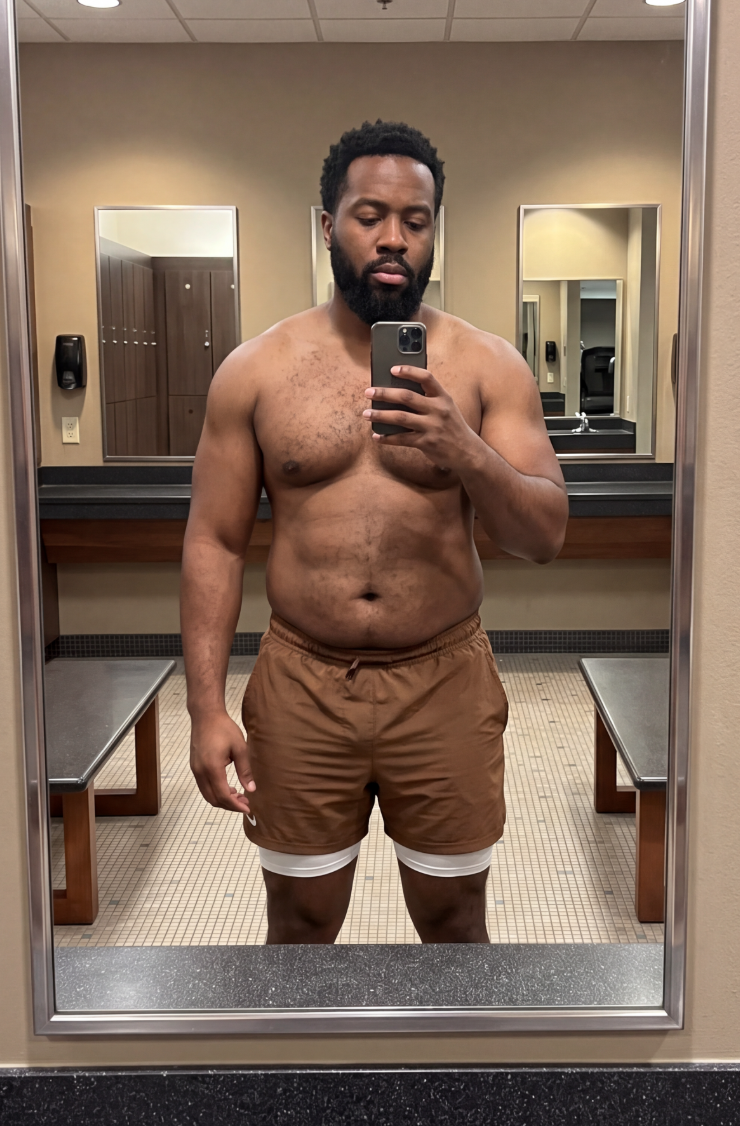 A shirtless man with a beard takes a selfie in a mirror inside a locker room.