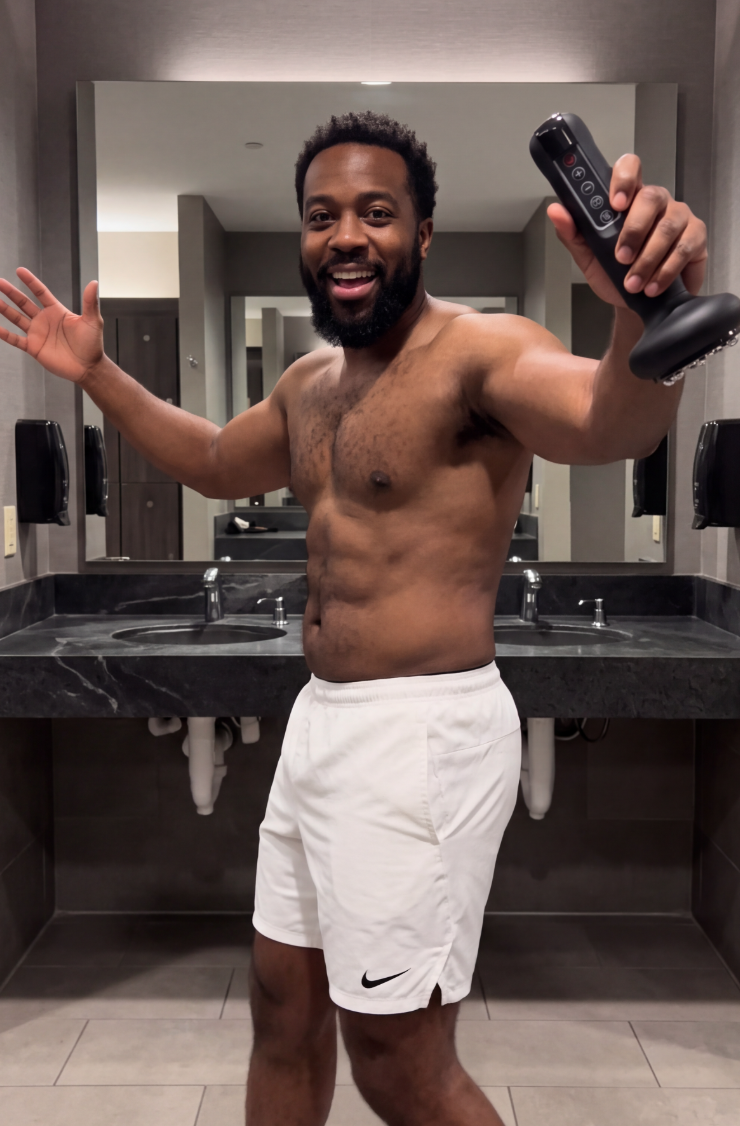 A smiling, shirtless man in white shorts holds up a black massage gun in a locker room.